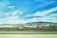 A bright red truck with a flatbed trailer is parked on a grassy area, with a backdrop of rolling hills and mountains under a blue sky with scattered clouds. A highway runs parallel in the foreground.