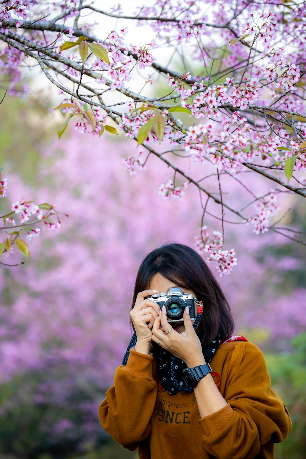 woman in orange jacket taking photo of pink cherry blossom