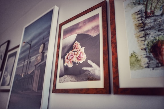 A series of framed pictures are hanging on a wall. The central image features a black and white photograph with a person's hand resting gently on a surface, adorned with light pink roses. The surrounding frames display various artworks, including what appears to be an urban scene and a landscape painting.