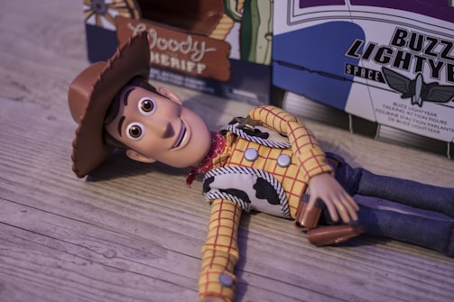 A toy figure dressed as a cowboy lies on a wooden surface. It wears a brown cowboy hat, yellow checkered shirt, red bandana, and blue jeans. A sheriff's badge is visible on its vest. In the background, partially visible boxes with logos related to space themes are present.