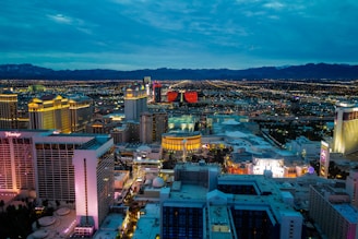 An extensive cityscape of Las Vegas is viewed from a high vantage point during dusk or early evening. The scene features numerous brightly lit hotels and casinos, displaying architectural variety. The sky is overcast with a hint of blue and the distant mountains provide a stark backdrop. The city lights form a glowing grid pattern that stretches toward the horizon.
