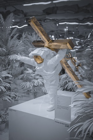 A white astronaut statue is depicted carrying a large, golden crucifix on its back. The astronaut stands on a pedestal surrounded by synthetic-looking plants. The setting appears to be an exhibition or gallery, given the presence of a sign that says 'PLEASE DO NOT TOUCH.' The overall atmosphere is surreal, with the combination of space and religious themes.