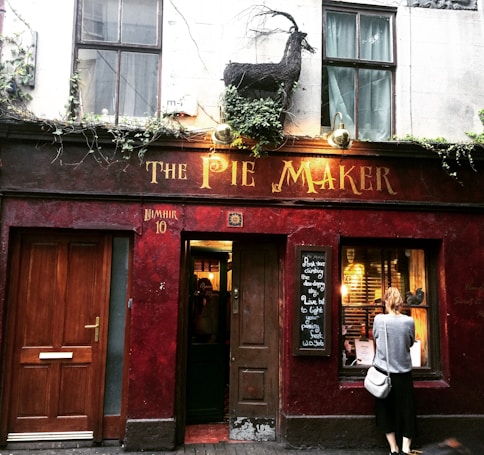 A rustic storefront with dark red walls features a sign reading 'The Pie Maker' in vintage-style lettering. Above the sign, a black goat sculpture and climbing plants add character. A person stands near a window looking at displayed items. Wooden doors and a chalkboard with handwritten text add to the cozy, inviting atmosphere.