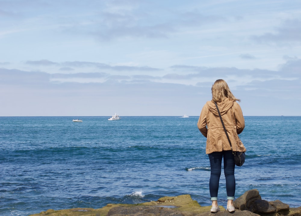 Frau in brauner Jacke tagsüber auf Felsen in der Nähe des Meeres