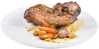 A piece of roasted meat seasoned with herbs, accompanied by roasted vegetables including carrots, garlic cloves, and a small onion on a white plate.