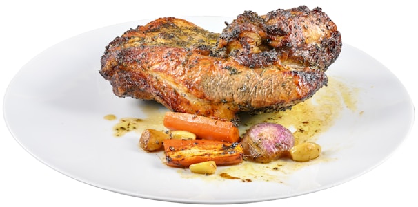 A piece of roasted meat seasoned with herbs, accompanied by roasted vegetables including carrots, garlic cloves, and a small onion on a white plate.