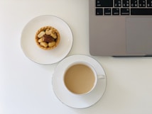 A neatly arranged scene features a cup of coffee with milk on a saucer, a small tart topped with various nuts on a plate, and the corner of a laptop on a clean white surface.