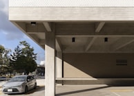 A modern concrete building with a minimalist design featuring large horizontal beams and vertical pillars. The building's exterior showcases a combination of smooth concrete surfaces and a portion of brickwork. A gray car is parked in front of the building, with several trees and a clear sky visible in the background.
