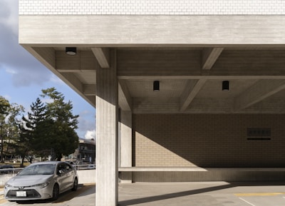 A modern concrete building with a minimalist design featuring large horizontal beams and vertical pillars. The building's exterior showcases a combination of smooth concrete surfaces and a portion of brickwork. A gray car is parked in front of the building, with several trees and a clear sky visible in the background.