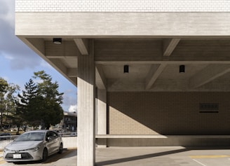A modern concrete building with a minimalist design featuring large horizontal beams and vertical pillars. The building's exterior showcases a combination of smooth concrete surfaces and a portion of brickwork. A gray car is parked in front of the building, with several trees and a clear sky visible in the background.