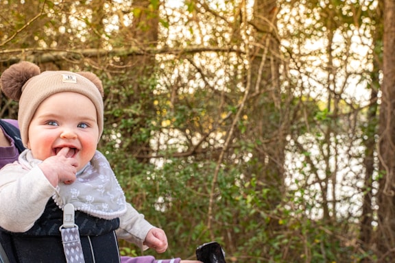 A baby wearing a beige knit hat with pom-poms and a patterned bib smiles with fingers in its mouth. The background is a natural setting with trees and foliage bathed in warm, golden light.