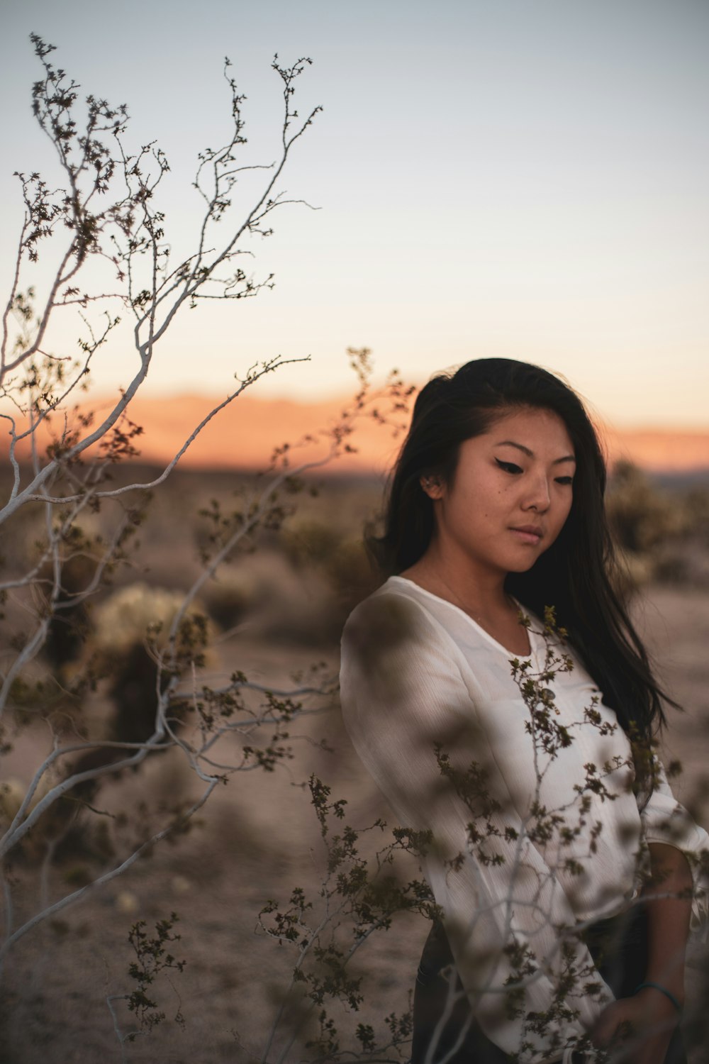 woman in white floral shirt standing near bare tree during sunset