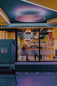 A nighttime scene of a small fast-food restaurant with a brightly lit interior, featuring a neon sign reading 'Lay's'. Two people sit inside at a table near the window, focused on their phones. The restaurant has a modern design with yellow and blue accents. Outside, sidewalks are visible with a barrier displaying some signage.