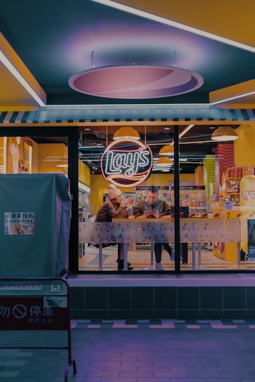 A nighttime scene of a small fast-food restaurant with a brightly lit interior, featuring a neon sign reading 'Lay's'. Two people sit inside at a table near the window, focused on their phones. The restaurant has a modern design with yellow and blue accents. Outside, sidewalks are visible with a barrier displaying some signage.