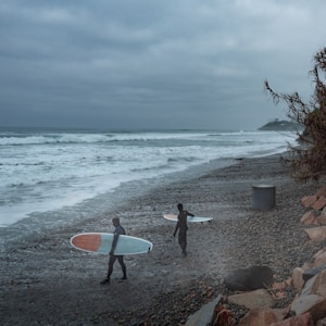 Two surfers in wetsuits walk along a pebbled beach carrying surfboards as they head towards the ocean. The waves crash lightly against the shore under a cloudy, overcast sky. A few plants and a rocky embankment line the right side of the scene, and a small headland can be seen in the distance.
