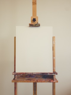 An empty, upright canvas sits on a wooden easel. The easel is spotted with different colors of paint, suggesting it has been used multiple times before. The background is a plain, solid color.