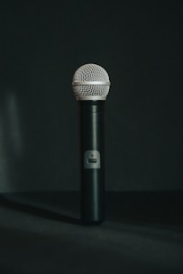 A black handheld microphone stands vertically against a dark background. The microphone has a metallic mesh grille at the top and a sleek black body with a small label or logo near the center.