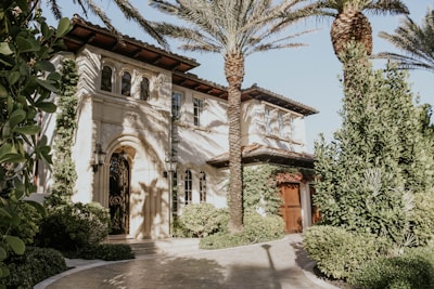 A Mediterranean-style house with stucco walls, arched windows, and a wrought iron gate. The property is surrounded by lush greenery, including palm trees and various shrubs. The sunlight creates intricate shadows on the facade, contributing to the serene atmosphere.