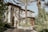A Mediterranean-style house with stucco walls, arched windows, and a wrought iron gate. The property is surrounded by lush greenery, including palm trees and various shrubs. The sunlight creates intricate shadows on the facade, contributing to the serene atmosphere.