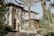 A Mediterranean-style house with stucco walls, arched windows, and a wrought iron gate. The property is surrounded by lush greenery, including palm trees and various shrubs. The sunlight creates intricate shadows on the facade, contributing to the serene atmosphere.
