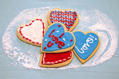 Five heart-shaped cookies decorated with colorful icing are arranged on a lightly dusted blue surface. The icing designs include red, white, and blue patterns, with the words 'love' and 'xoxo' written on two of the cookies in a playful font.