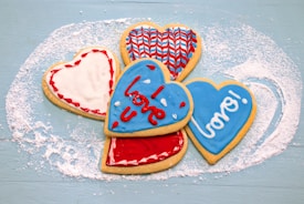 Five heart-shaped cookies decorated with colorful icing are arranged on a lightly dusted blue surface. The icing designs include red, white, and blue patterns, with the words 'love' and 'xoxo' written on two of the cookies in a playful font.