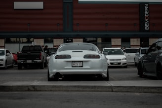 A parking lot scene features a variety of cars, with a white sports car prominently in the center, displaying a distinctive license plate. A modern building with horizontal lines and signage forms the backdrop, adding a sense of urban setting. Several parked vehicles of different makes and colors are visible, and a few people can be seen in the background.