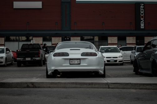 A parking lot scene features a variety of cars, with a white sports car prominently in the center, displaying a distinctive license plate. A modern building with horizontal lines and signage forms the backdrop, adding a sense of urban setting. Several parked vehicles of different makes and colors are visible, and a few people can be seen in the background.