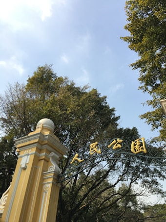 An ornate entrance gate with gold lettering stands prominently against a backdrop of lush green trees. The structure features classic architectural elements with yellow and white accents, complemented by a clear blue sky overhead.