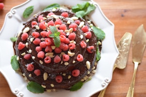 A chocolate cake is adorned with fresh raspberries, white chocolate chips, and mint leaves. It is displayed on a decorative white plate. The cake is rich and glossy, with the texture and sheen of the chocolate icing clearly visible. Around the cake, there are golden utensils placed on a wooden surface.