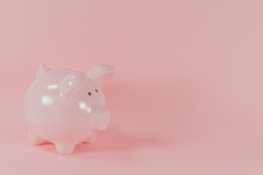A light pink ceramic piggy bank is placed on a solid pink background. The surface of the piggy bank is glossy and it has small black eyes.