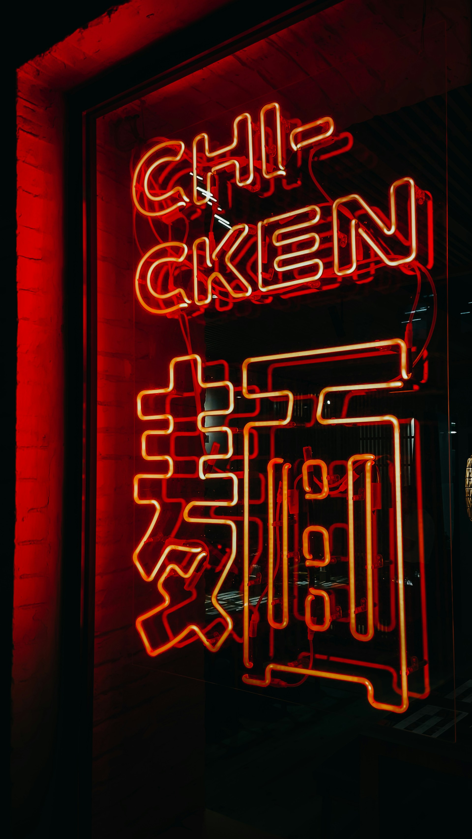 A brightly glowing neon sign with English and possibly Chinese characters. The word 'CHICKEN' is prominently displayed in red neon light. Below it, a series of neon characters add an additional cultural layer to the sign. The neon sign is placed in a window and casts a red glow on the surroundings.