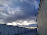 A row of large, white storage containers is aligned along a gravel road. The sky overhead is filled with dark, dramatic clouds, with patches of pale blue and hints of sunset on the horizon. An industrial pole stands near the containers, and a vehicle is partially visible along the side.