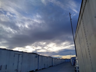 A row of large, white storage containers is aligned along a gravel road. The sky overhead is filled with dark, dramatic clouds, with patches of pale blue and hints of sunset on the horizon. An industrial pole stands near the containers, and a vehicle is partially visible along the side.
