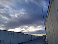A row of large, white storage containers is aligned along a gravel road. The sky overhead is filled with dark, dramatic clouds, with patches of pale blue and hints of sunset on the horizon. An industrial pole stands near the containers, and a vehicle is partially visible along the side.