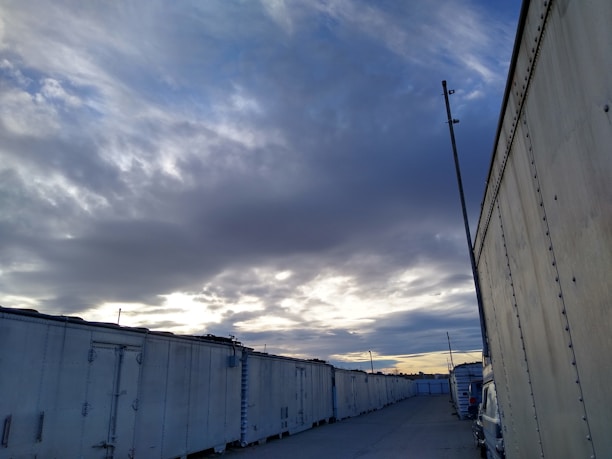 A row of large, white storage containers is aligned along a gravel road. The sky overhead is filled with dark, dramatic clouds, with patches of pale blue and hints of sunset on the horizon. An industrial pole stands near the containers, and a vehicle is partially visible along the side.