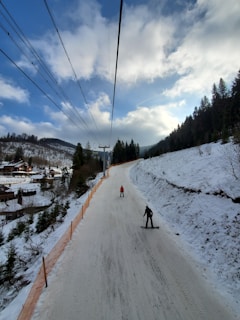 A snowy landscape with a ski slope surrounded by forested hills under a partly cloudy blue sky. Two people are skiing downhill, with a ski lift stretching above them. There are wooden cabins and trees along the left, indicating a resort area.