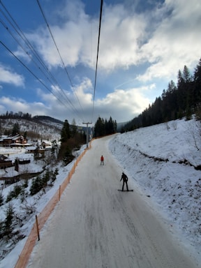 A snowy landscape with a ski slope surrounded by forested hills under a partly cloudy blue sky. Two people are skiing downhill, with a ski lift stretching above them. There are wooden cabins and trees along the left, indicating a resort area.