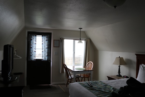 A small, cozy room with a slanted ceiling features a bed, a small table with two wooden chairs near a window, and a door with patterned curtains. A lamp sits on a nightstand next to the bed, and a TV is partially visible on the left.
