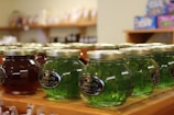 Several jars of pepper jelly are arranged on a wooden shelf, with labels indicating jalapeño flavor. The jars have metallic lids and come in various colors, like green and red. In the background, other shelves and products are slightly blurred.