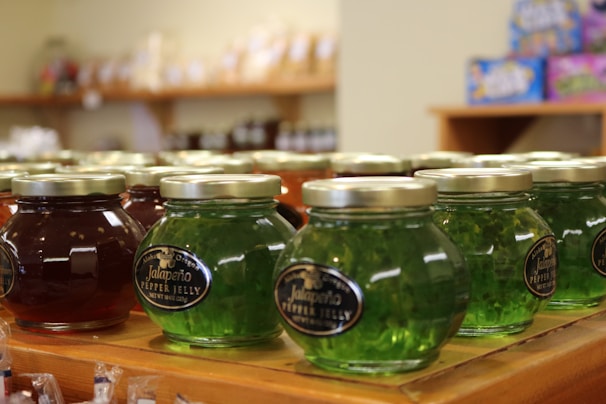 Several jars of pepper jelly are arranged on a wooden shelf, with labels indicating jalapeño flavor. The jars have metallic lids and come in various colors, like green and red. In the background, other shelves and products are slightly blurred.