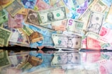 A collection of various banknotes from different countries is scattered across a reflective surface. The currencies include US dollars, Mexican pesos, and Indian rupees, among others. The banknotes feature diverse colors and designs, with some displaying portraits and others showcasing intricate patterns.
