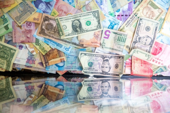 A collection of various banknotes from different countries is scattered across a reflective surface. The currencies include US dollars, Mexican pesos, and Indian rupees, among others. The banknotes feature diverse colors and designs, with some displaying portraits and others showcasing intricate patterns.