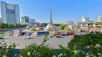 A busy urban roundabout with a tall central monument surrounded by traffic. Cars and buses are making their way around the circular road, and there are large buildings and billboards in the background. Trees and greenery are present in the foreground.