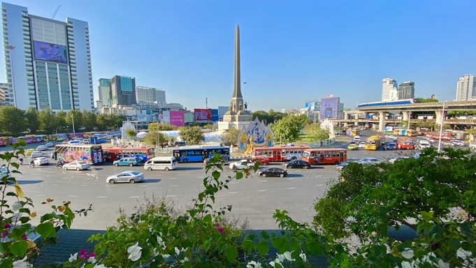 A busy urban roundabout with a tall central monument surrounded by traffic. Cars and buses are making their way around the circular road, and there are large buildings and billboards in the background. Trees and greenery are present in the foreground.