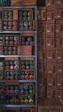 Shelves filled with assorted spray paint cans of various colors, including red, green, orange, and yellow, alongside unopened cardboard boxes. The boxes are stacked neatly next to the shelves, labeled 'COLORART' and 'NOU COLORS'.