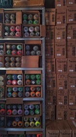 Shelves filled with assorted spray paint cans of various colors, including red, green, orange, and yellow, alongside unopened cardboard boxes. The boxes are stacked neatly next to the shelves, labeled 'COLORART' and 'NOU COLORS'.