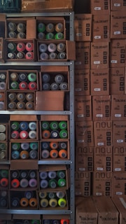 Shelves filled with assorted spray paint cans of various colors, including red, green, orange, and yellow, alongside unopened cardboard boxes. The boxes are stacked neatly next to the shelves, labeled 'COLORART' and 'NOU COLORS'.