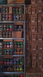 Shelves filled with assorted spray paint cans of various colors, including red, green, orange, and yellow, alongside unopened cardboard boxes. The boxes are stacked neatly next to the shelves, labeled 'COLORART' and 'NOU COLORS'.