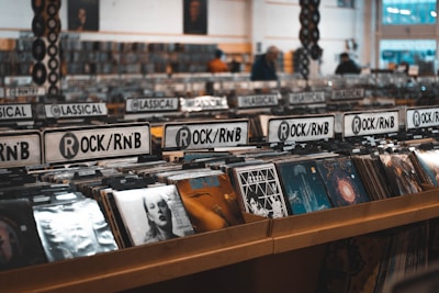 Rows of vinyl records are displayed in a music store, categorized under various genres such as Rock/RnB and Classical. The shelves are filled with album covers featuring diverse artwork. The space is well-lit, suggesting an inviting atmosphere for browsing music enthusiasts.